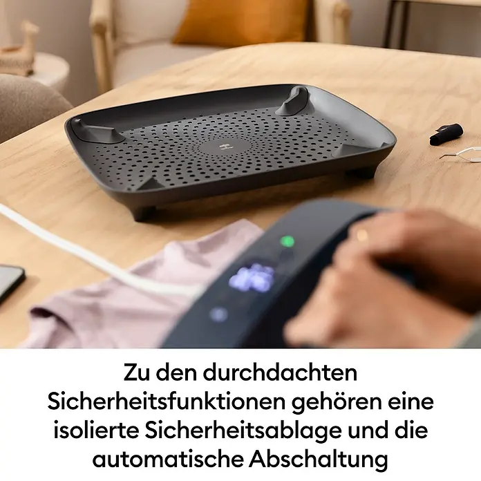 Cricut Transferpresse EasyPress SE + Hitzeschutzmatte - 25 x 30 cm, Passend für: Bügelfolien - Dunkelgraue Sicherheitsablage mit perforierter Oberfläche auf einem Holztisch, im Vordergrund dämpft eine Hand ein rosa Kleidungsstück; zeigt durchdachte Sicherheitsfunktionen.