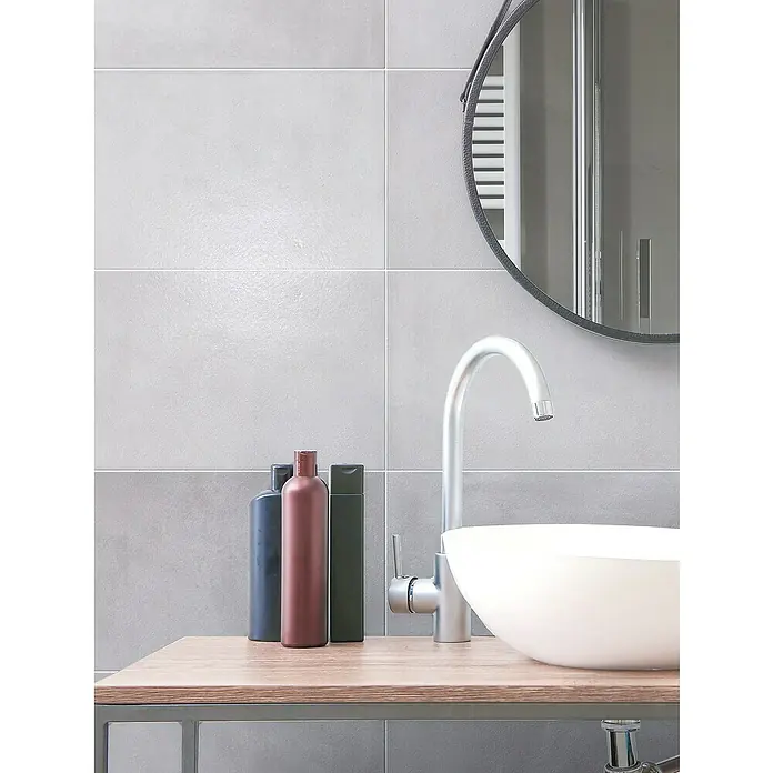 Steingutfliese Flower - 60 x 30 cm, Light Grey, Matt - Modernes Badezimmer mit weißem Aufsatzwaschbecken, silbernem Wasserhahn und drei farbigen Flaschen auf Holzplatte vor grauen Fliesen.