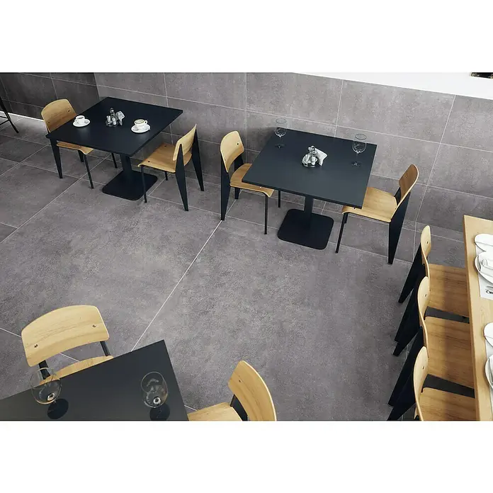 Feinsteinzeugfliese Savanna - 59,7 x 119,7 cm, Anthrazit, Matt - Modernes Café-Interieur mit großformatigen grauen Betonoptik-Fliesen an Boden und Wänden, ausgestattet mit schwarzen Tischen und Holzstühlen.