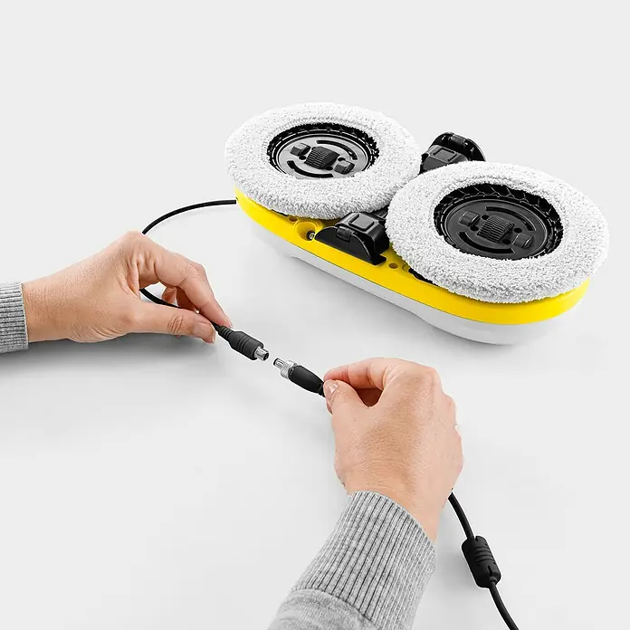 Kärcher Fensterreinigungsroboter RCW 2 - Wassertankvolumen Roboter: 70 ml, Saugkraft: 3.300 Pa - Kärcher Bodenreiniger in Gelb und Weiß mit weißen Walzen, Hände verbinden das schwarze Ladekabel.