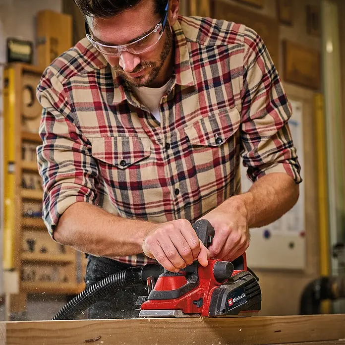 Einhell Elektrohobel TE-PL 920 - 920 W - Mann in Karohemd verwendet roten und schwarzen Einhell Elektrohobel zum Glätten von Holz, Späne fallen.