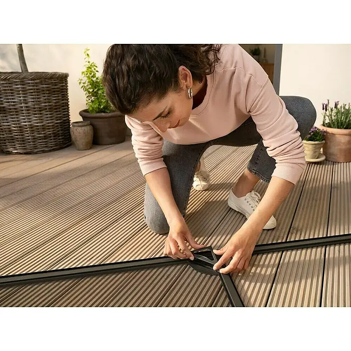 Tesa Insektenschutztür - B x H: 1,2 x 2,4 m, Weiß, Montage Insektengitter: Schrauben - Eine Person montiert ein Eckstück eines schwarzgerahmten Insektenschutzgitters mit dunklem Netz auf einer Holzterrasse.