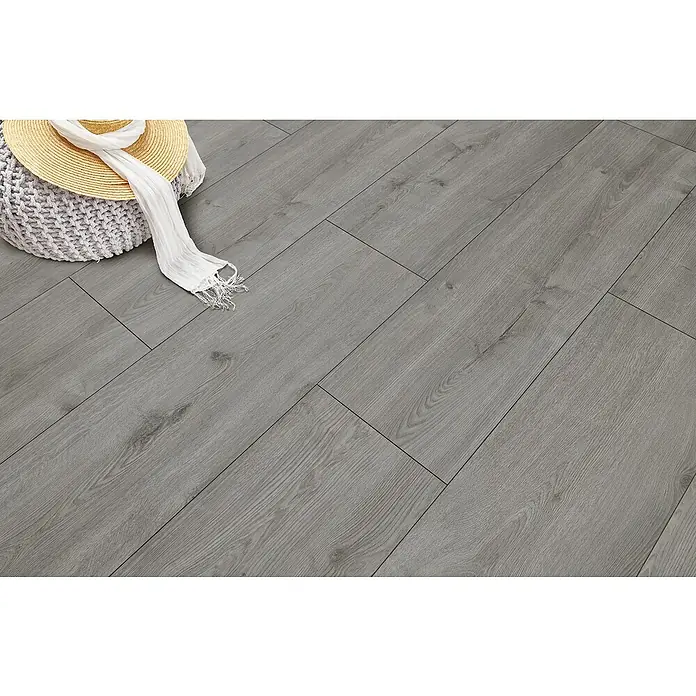 LOGOCLIC Vinto Laminat - Eiche Calais, Landhausdiele, 1.288 x 330 x 8 mm - Grauer Laminatboden in Holzoptik. Ein Hut und Schal liegen auf einem Pouf.