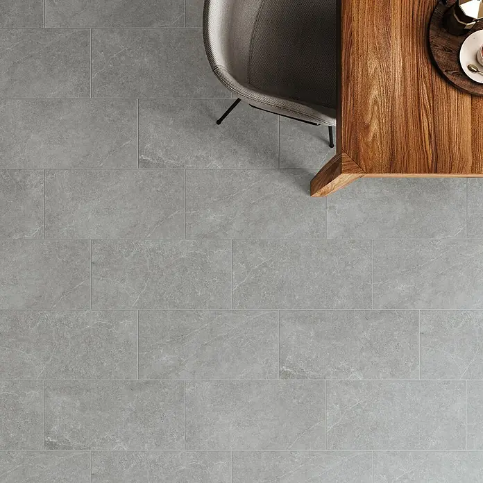 DryTile Feinsteinzeugfliese Silkstone Schlamm - 30 x 60 cm, Grau, Matt - Graue rechteckige Fliesen mit subtiler Steintextur als Bodenbelag, neben einem Holztisch und Stuhl.
