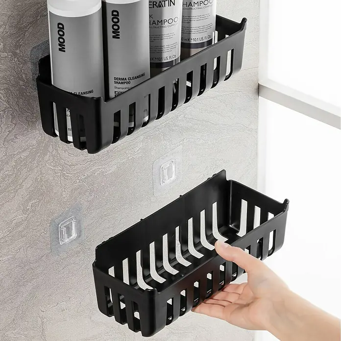 Cesta de baño Molly - 11 x 25 x 9 cm, Plástico, Negro - Dos cestas de ducha de plástico negro, una montada con ganchos adhesivos transparentes en una pared de azulejos, la otra sostenida por una mano.
