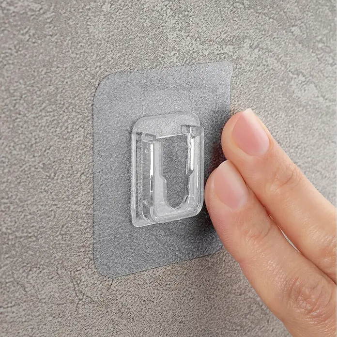Cesta de baño Molly - 11 x 25 x 9 cm, Plástico, Negro - Clip de pared autoadhesivo transparente siendo presionado con los dedos sobre una superficie texturizada. Ideal para organizar.