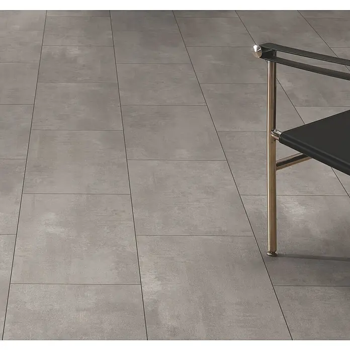 Laminat - Oxyd, Fliese, 1.288 x 330 x 8 mm - Großformatige graue Betonoptik-Bodenfliesen, teilweise sichtbar mit einem modernen Stuhl.