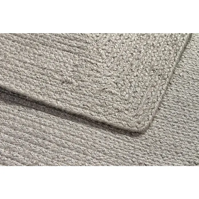 Alfombra exterior In/Out Ebro - Plata, 110 x 60 cm, 100% Polietileno - Primer plano de una alfombra textil trenzada gris claro, mostrando textura intrincada y una esquina doblada.