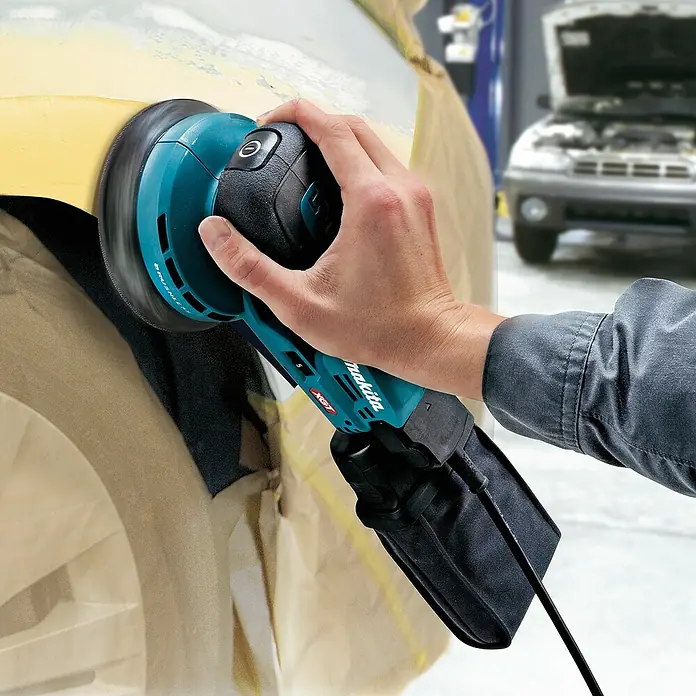 Makita XGT 40V max. Akku-Exzenterschleifer - 40 V, Ohne Akku, Durchmesser Schleifteller: 125 mm - Eine Hand hält einen türkis-schwarzen Makita Exzenterschleifer, der ein Autoteil schleift.