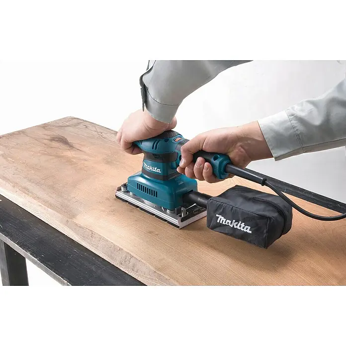 Makita Schwingschleifer BO3711J - 190 W, Länge Schleifplatte: 185 mm, Breite Schleifplatte: 93 mm - Person schleift Holz mit einem türkisfarbenen und schwarzen Makita Schwingschleifer mit Staubbeutel.