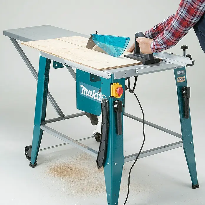 Makita Tischkreissäge 2712 - 2.000 W, Durchmesser Sägeblatt: 315 mm, Schnitttiefe bei 90 °: 85 mm - Türkise Makita Tischsäge schneidet helle Holzbretter, eine Person bedient sie. Sägemehl liegt um die Basis.