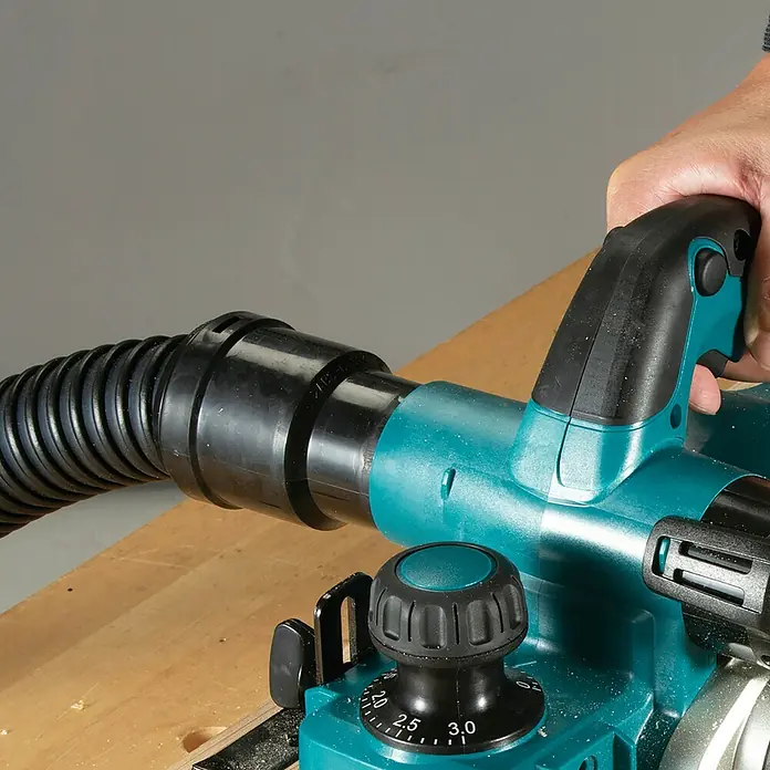 Makita LXT 18V Akku-Hobel - 18 V, Ohne Akku, Leerlaufdrehzahl: 12.000 U/min - Hand hält einen türkis-schwarzen Elektrohobel mit angeschlossenem Staubschlauch auf einer Holzoberfläche.