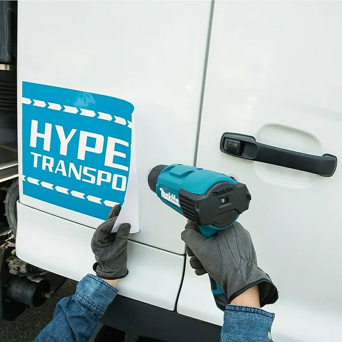 Makita Heißluftgebläse HG6031VK - 1.800 W, Temperaturbereich: 50 °C - 600 °C - Behandschuhte Hände verwenden eine türkis-schwarze Makita Heißluftpistole, um ein 'HYPE TRANSPO' Abziehbild auf ein weißes Fahrzeug anzubringen.
