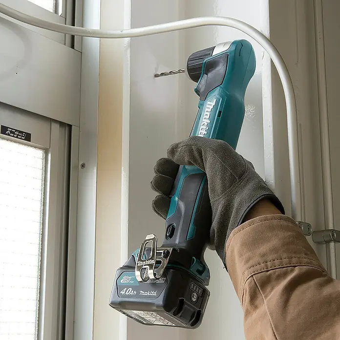 Makita CXT 12V max. Akku-Winkelbohrmaschine DA333DZ - 12 V, Ohne Akku - Eine Person bohrt mit einem türkis-schwarzen Makita Akku-Winkelbohrer ein Loch in eine weiße Wand, nahe Fenster und Metallrohr.