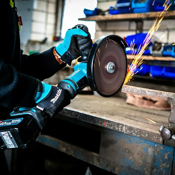 Makita XGT 40V max. Akku-Winkelschleifer GA038GZ - Ohne Akku, Durchmesser Scheibe: 230 mm - Makita Winkelschleifer (türkis-schwarz) schneidet Metallstange, erzeugt Funken, gehalten von Händen mit passenden Handschuhen.