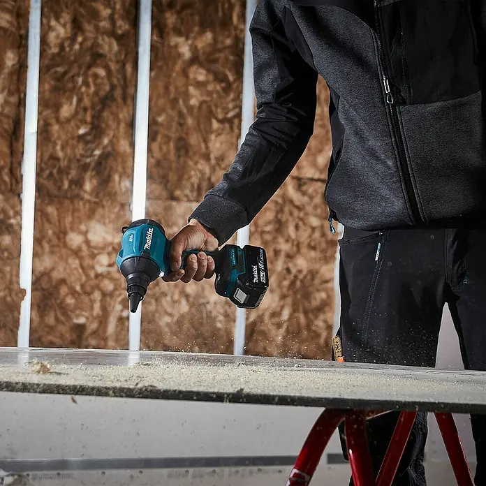 Makita LXT 18V Akku-Gebläse DAS180Z - 18 V, Ohne Akku, 200 mm/s - Nahaufnahme einer Person, die einen türkis-schwarzen Makita Akku-Schlagschrauber aktiv auf einer Oberfläche benutzt und feinen Sägemehl erzeugt.