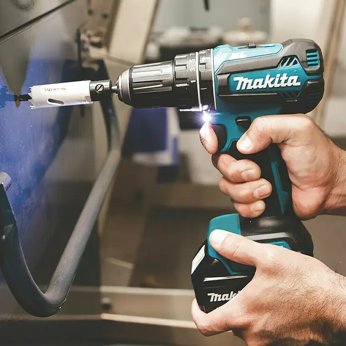 Makita LXT 18V Akku-Schlagschrauber DHP485Z - 18 V, Ohne Akku, 50 Nm - Eine Person hält einen teal-schwarzen Makita Akkubohrschrauber mit leuchtender LED, der aktiv mit einer weißen Lochsäge in eine blau-graue Oberfläche bohrt.