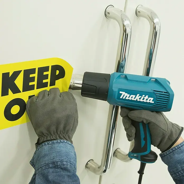 Makita Heißluftgebläse HG5030K - 1.600 W, Temperaturbereich: 350 °C - 500 °C - Eine Person entfernt mit einer blaugrünen und schwarzen Makita Heißluftpistole ein gelbes Schild.