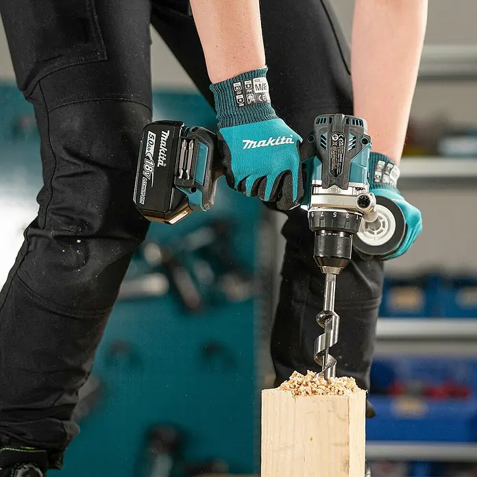 Makita LXT 18V Akku-Schlagbohrschrauber DHP492RTJ - 18 V, 2 Akkus, 5 Ah, 130 Nm - Eine Person bohrt mit einem teal-schwarzen Makita Akku-Bohrschrauber mit 18V Li-ion Akku in Holz. Späne fliegen vom großen Holzbohrer.