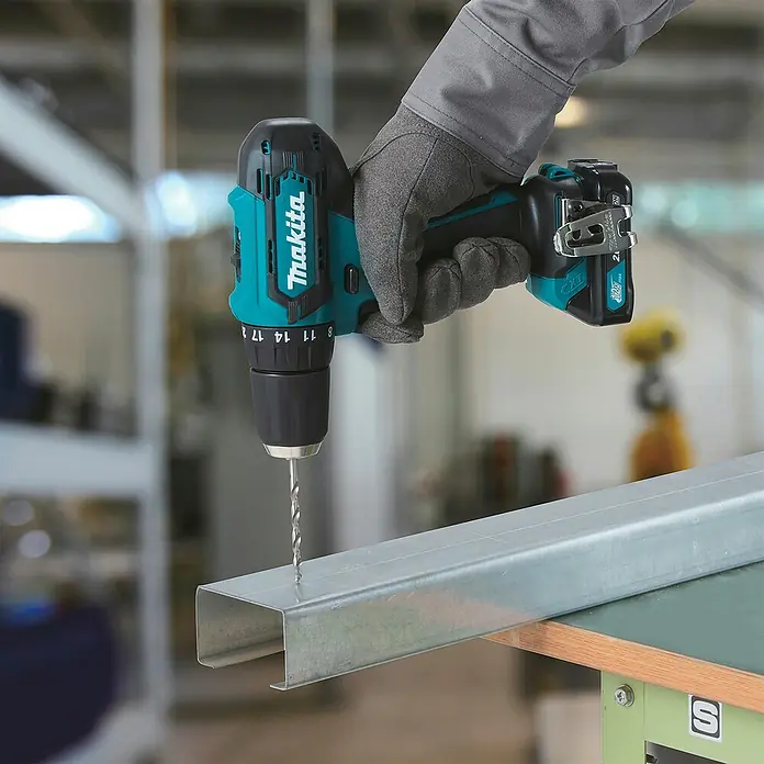 Makita CXT 12V max. Akku-Schlagbohrschrauber HP333DZ - 12 V, Ohne Akku, 30 Nm, Ohne Akku und Zubehör - Eine behandschuhte Hand hält einen türkis-schwarzen Makita Akkubohrschrauber, der in ein silbernes Metallprofil bohrt.