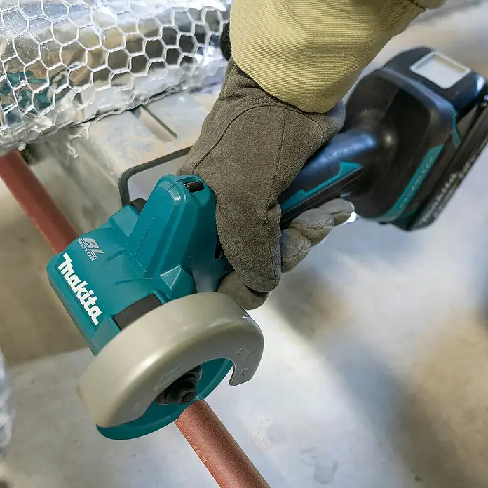 Makita LXT 18V Akku-Trennschleifer DMC300Z - Ohne Akku - Behandschuhte Hand benutzt eine türkis-schwarze Makita Akku-Trennsäge zum Schneiden eines Kupferrohrs.