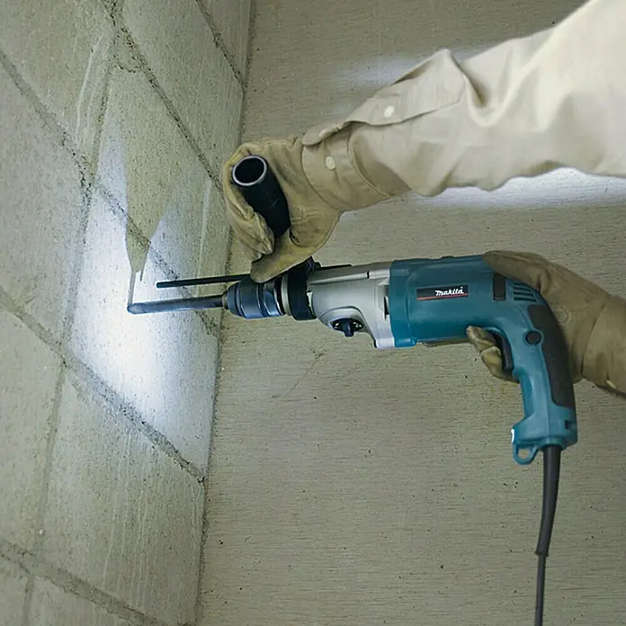 Makita Schlagbohrmaschine HP2071J - 1.100 W - Person in Handschuhen bedient einen blau-grauen Schlagbohrer, der in eine Betonwand bohrt; heller Lichtpunkt am Bohrpunkt.