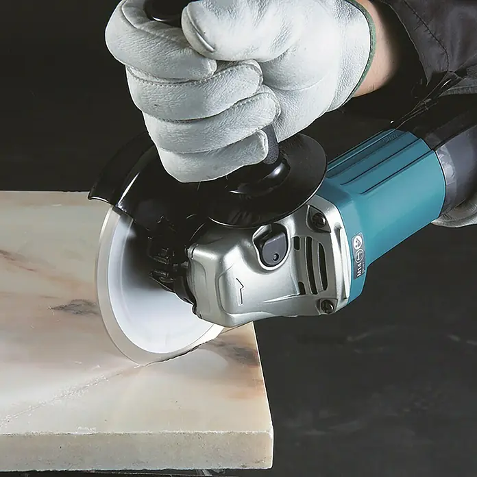 Makita Elektro-Winkelschleifer GA5030R - 720 W, 11.000 U/min - Winkelschleifer mit weißer Trennscheibe, von behandschuhter Hand gehalten, schneidet in helle Steinplatte.