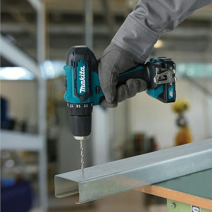 Makita CXT 12V max. Akku-Bohrschrauber DF333DZ - 12 V, Ohne Akku - Eine behandschuhte Hand hält einen türkisen und schwarzen Makita Akkubohrer, der ein Loch in ein silbernes Metall-C-Profil bohrt.