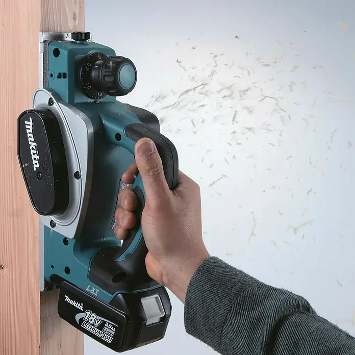 Makita LXT 18V Akku-Handhobel DKP180Z - 18 V, Li-Ionen, Ohne Akku, Hobelbreite: 82 mm - Ein blaugrüner und silberner Makita Akku-Hobel wird von einer Hand gehalten, die einen Holzbalken hobelt, mit Holzspänen, die herausfliegen. Ein 18V Lithium-Ionen-Akku ist angebracht.