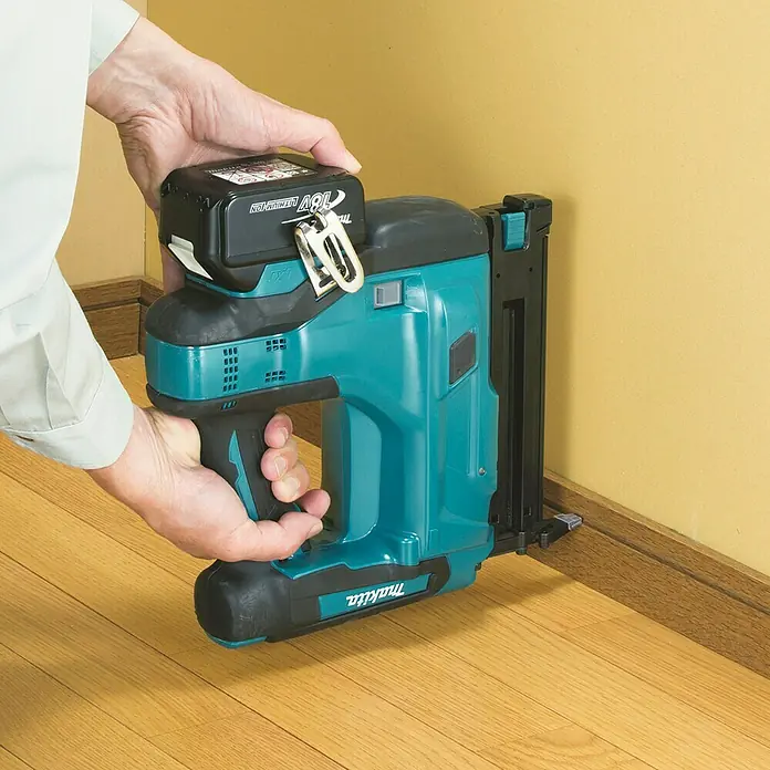 Makita LXT 18V Akku-Nagler DBN500RTJ - 18 V, 2 Akkus, 5 Ah, Länge Nagel: 15 mm - 50 mm - Person benutzt eine türkis-schwarze Makita Akku-Nagelpistole mit 18V Akku zum Befestigen einer Fußleiste an gelber Wand.