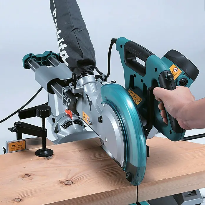 Makita Kapp- & Gehrungssäge LS1018LN - 1.430 W, Durchmesser Sägeblatt: 260 mm, 4.300 U/min - Türkis-graue Gehrungssäge mit Staubfangbeutel und transparentem Blattschutz, schneidet einen Holzbalken. Eine Hand bedient die Säge.