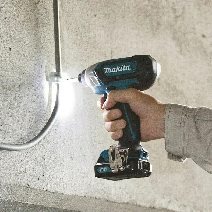 Makita Akku-Schlagschrauber TD110DSMJ - 10,8 V, 2 Akkus, 4 Ah - Hand hält einen schwarz-türkisen Makita Schlagschrauber, dessen Licht eine Schraube an einer Wand neben einem Rohr beleuchtet.