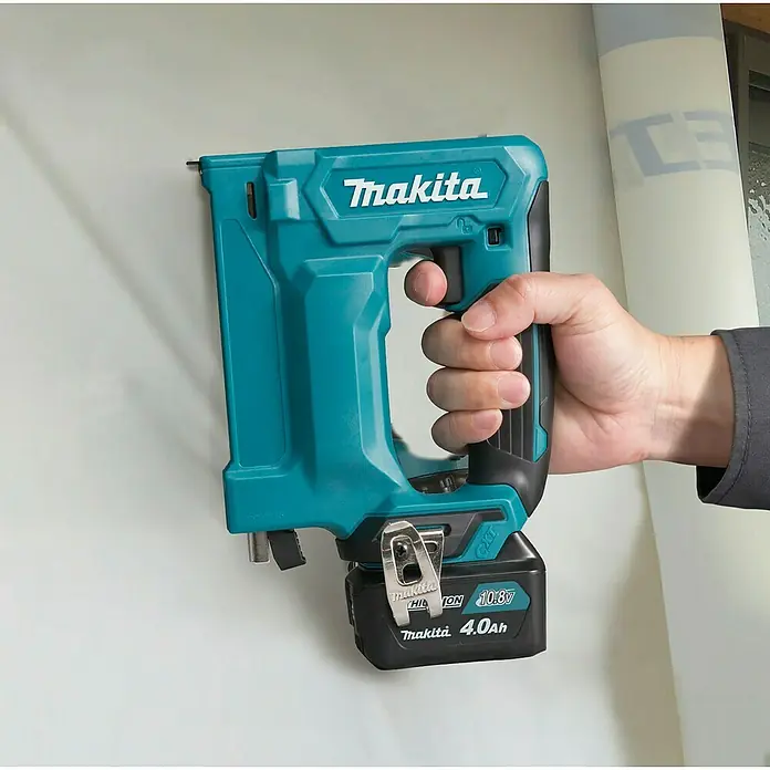 Makita CXT 12V max. Akku-Tacker ST113DSMJ - 12 V, 2 Akkus, 4 Ah - Eine Hand hält eine türkis-schwarze Makita Akku-Klammerpistole und heftet helles Material an eine Wand.