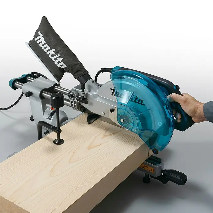 Makita Kapp- & Gehrungssäge LS1110F - 1.400 W, Durchmesser Sägeblatt: 260 mm, Leerlaufdrehzahl: 4.500 U/min - Makita Kapp- und Gehrungssäge mit blauem Sägeblattschutz und schwarzem Staubsack, sägt ein helles Holzbrett.