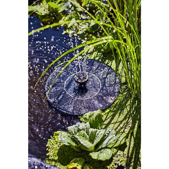 Heissner Zierbrunnen Solar Wassergarten Lotusgarden - Anthrazit, L x B x H: 58 x 58 x 19 cm - Schwarze, runde Solar-Springbrunnenpumpe schwimmt in Wasser, sprüht Wassertropfen, umgeben von grünen Pflanzen.