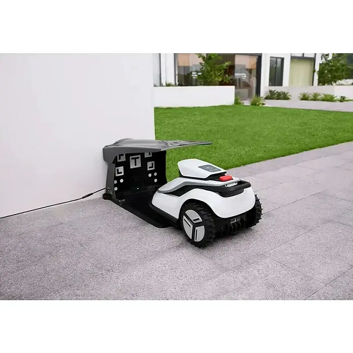 Terramow Mähroboter-Set V600 inkl. Garage - 600 m², Schnittbreite: 20,3 cm, Kamera-gestützte Navigation - Weißer und schwarzer Rasenmähroboter mit Profilrädern, angedockt in seiner schwarzen Ladestation mit grauem Dach.