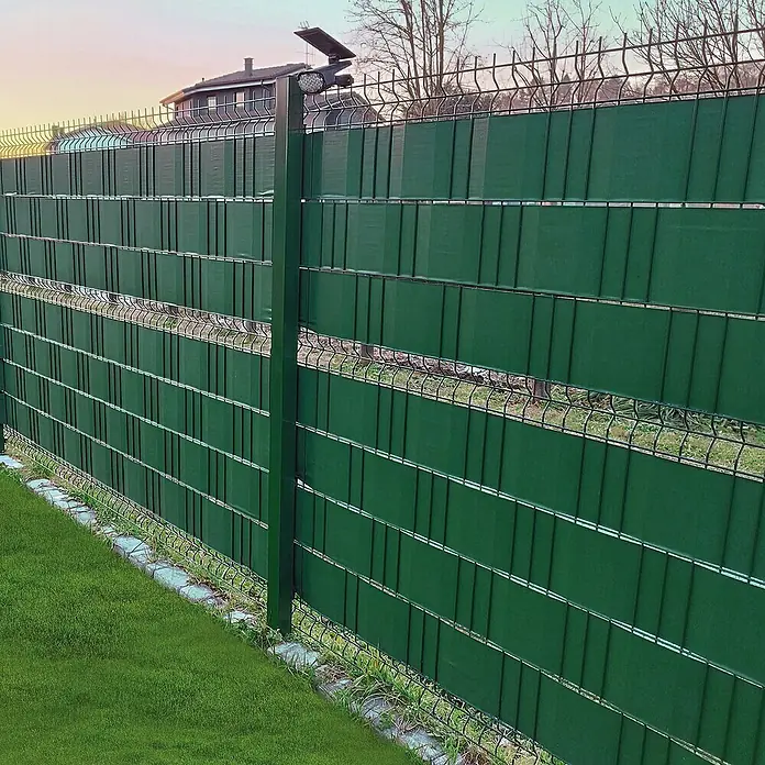 Tenax Malla de ocultación Twist - 35 x 0,19 m, Verde - Valla de malla metálica verde con tiras de privacidad y luz solar, instalada en un jardín.