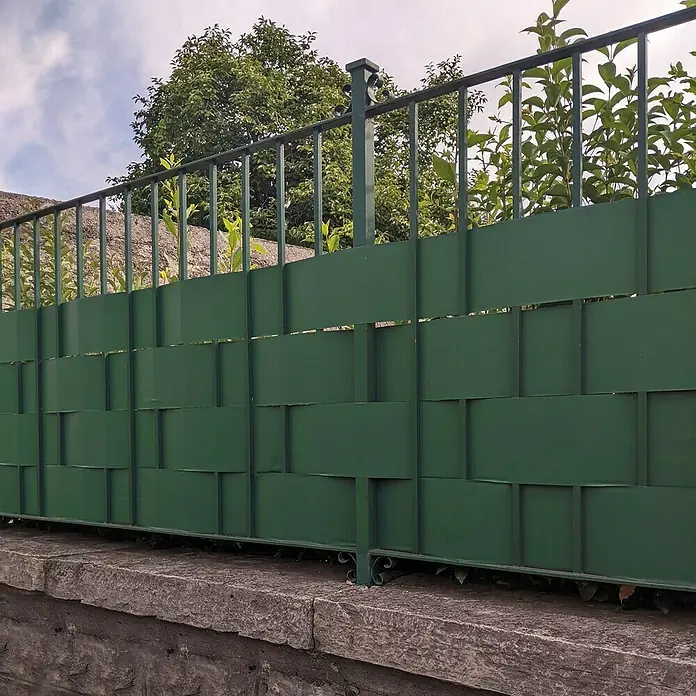 Tenax Malla de ocultación Twist - 35 x 0,19 m, Verde - Valla de privacidad verde tejida sobre un muro de piedra, con plantas visibles arriba.