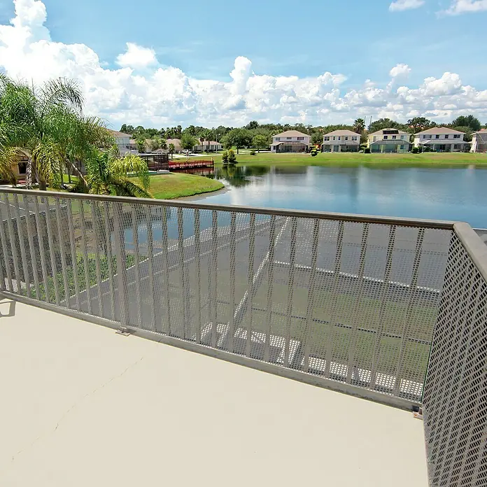 Tenax Malla protectora Quadra 10 - L x Al: 5 x 1 m, Plata - Barandilla de metal perforado gris en un balcón con vistas a un lago y casas.