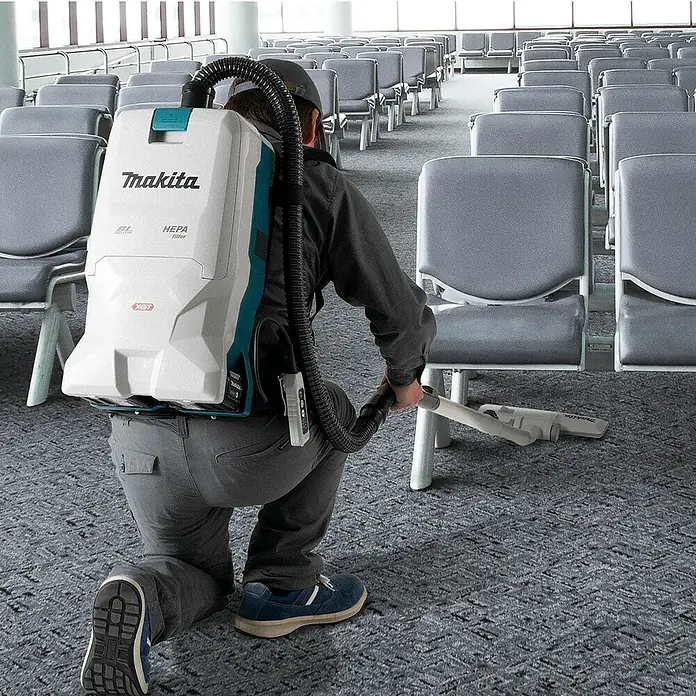 Makita XGT 40V max. Aspiradora con mochila de batería VC011GZ - 40 V, Sin batería, Volumen de recipiente: 6 l - Aspiradora de mochila Makita blanca/turquesa, usada por persona arrodillada para limpiar pasillo alfombrado entre sillas.