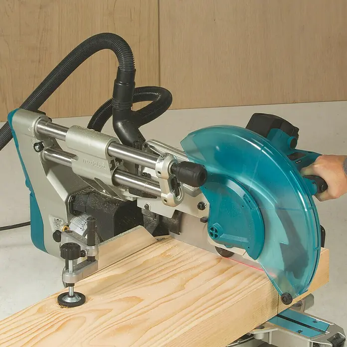 Makita Kapp- & Gehrungssäge LS1219L - 1.800 W, Durchmesser Sägeblatt: 305 mm, Leerlaufdrehzahl: 0 U/min - 3.200 U/min - Türkis-silberne Kappsäge mit transparentem Blattschutz sägt ein Holzbrett. Staubsaugerschlauch und Laserführung sind sichtbar.