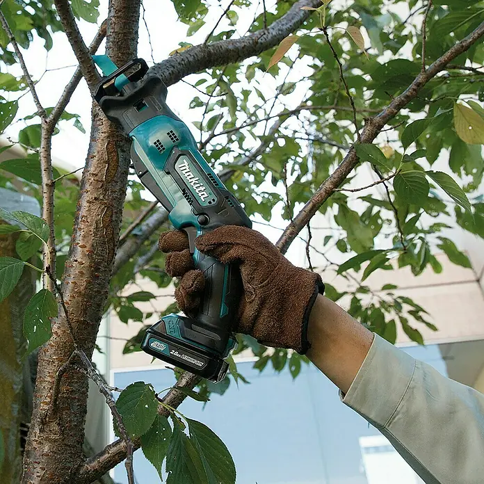 Makita Akku-Säbelsäge JR103DZ - 10,8 V, Ohne Akku, Leerlaufhubzahl: 0 /min - 3.300 /min - Makita Akku-Reciprosäge in Türkis und Schwarz, von einer behandschuhten Hand gehalten, beim Schneiden eines Astes.