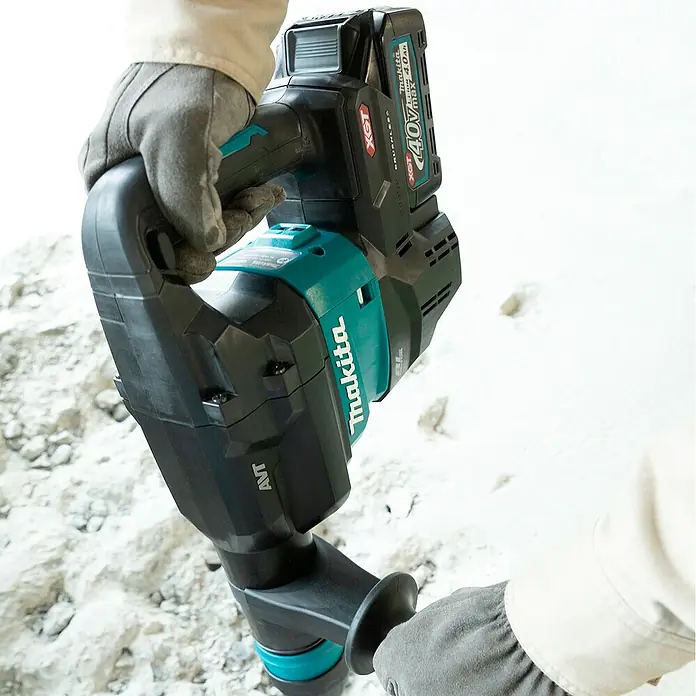 Makita XGT 40V max. Martillo combinado de batería HM001G - 40 V, Sin batería, 9,4 J - Una persona con guantes opera un taladro percutor inalámbrico Makita negro y turquesa con batería de 40V, perforando una superficie clara.