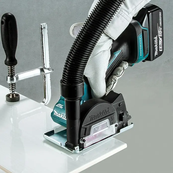 Makita LXT 18V Akku-Trennschleifer DMC300Z - Ohne Akku - Eine behandschuhte Hand bedient einen schwarzen und türkisfarbenen Makita Akku-Fliesenschneider mit Absaugschlauch, der eine mit einer Klemme gesicherte weiße Fliese schneidet.
