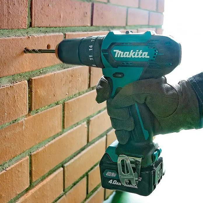 Makita CXT 12V max. Akku-Schlagbohrschrauber HP333DZ - 12 V, Ohne Akku, 30 Nm, Ohne Akku und Zubehör - Eine Person mit Handschuhen verwendet einen türkisfarbenen und schwarzen Makita 10.8V Akku-Bohrschrauber mit 4.0Ah Akku, um ein Loch in eine Ziegelwand zu bohren.