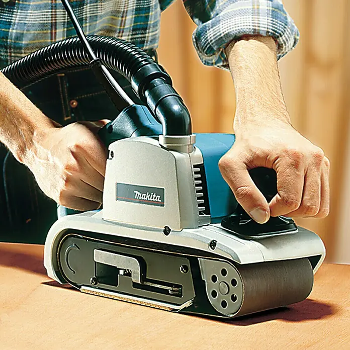 Makita Elektro-Bandschleifmaschine 9403J - 1.200 W - Eine Person schleift eine Holzoberfläche mit einem silber-blauen Makita Bandschleifer, der an einen schwarzen Absaugschlauch angeschlossen ist.