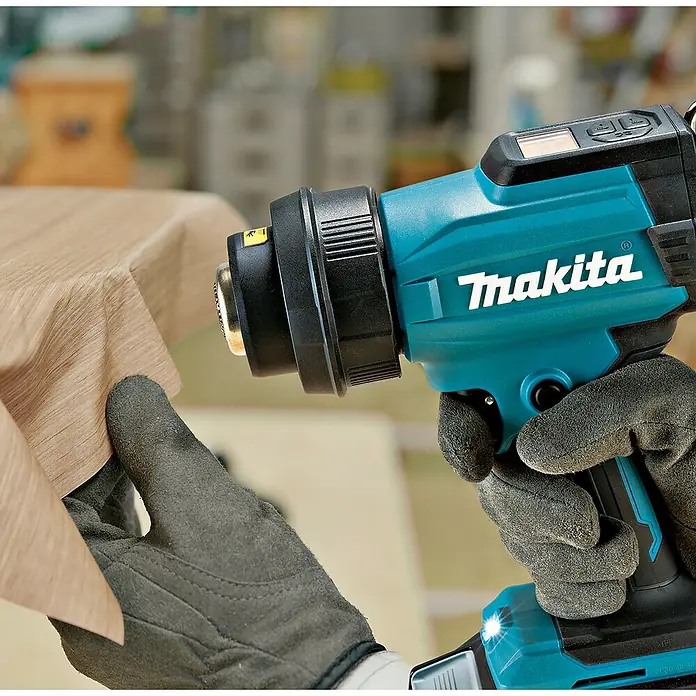 Makita XGT 40V max. Akku-Heißluftgebläse HG001G - 40 V, Ohne Akku, Temperaturbereich: 0 °C - 600 °C - Behandschuhte Hände halten eine türkis-schwarze Makita Heißluftpistole, die Wärme zum Formen oder Anbringen von hellem Holzkantenband anwendet.