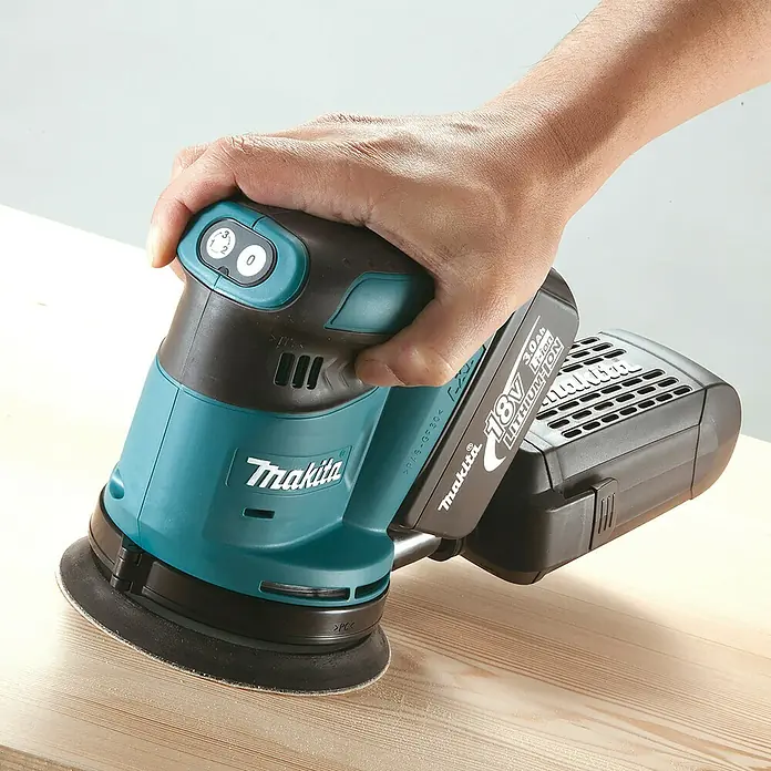 Makita LXT 18V Lijadora excéntrica de batería DBO180RTJ -  - Una mano opera una lijadora orbital inalámbrica Makita 18V turquesa y negra sobre madera clara.