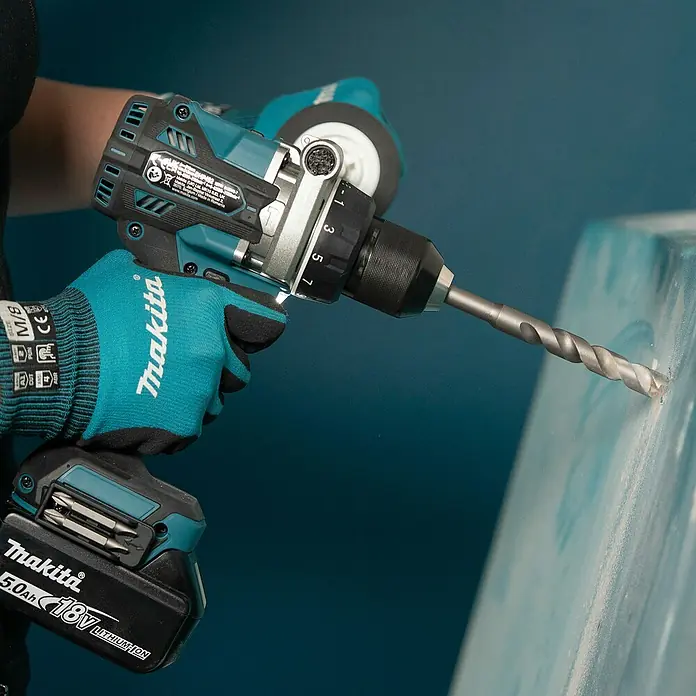 Makita LXT 18V Akku-Schlagbohrschrauber DHP492RTJ - 18 V, 2 Akkus, 5 Ah, 130 Nm - Eine Person mit Makita-Handschuhen hält eine türkis-schwarze Makita 18V Akkubohrmaschine, die in eine hellblaue Oberfläche bohrt.