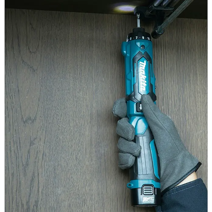 Makita Akkuschrauber DF012DSE - 7,2 V, 2 Akkus, 1,5 Ah - Türkisfarbener und schwarzer Makita Akkuschrauber, von einer behandschuhten Hand unter einem Holzschrank benutzt, die Arbeitsfläche beleuchtend.